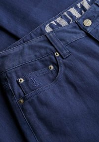 Vaqueros de mezclilla azul oscuro con un bolsillo frontal con remaches plateados, un logotipo bordado y una cinturilla marcada con letras blancas desvanecidas.