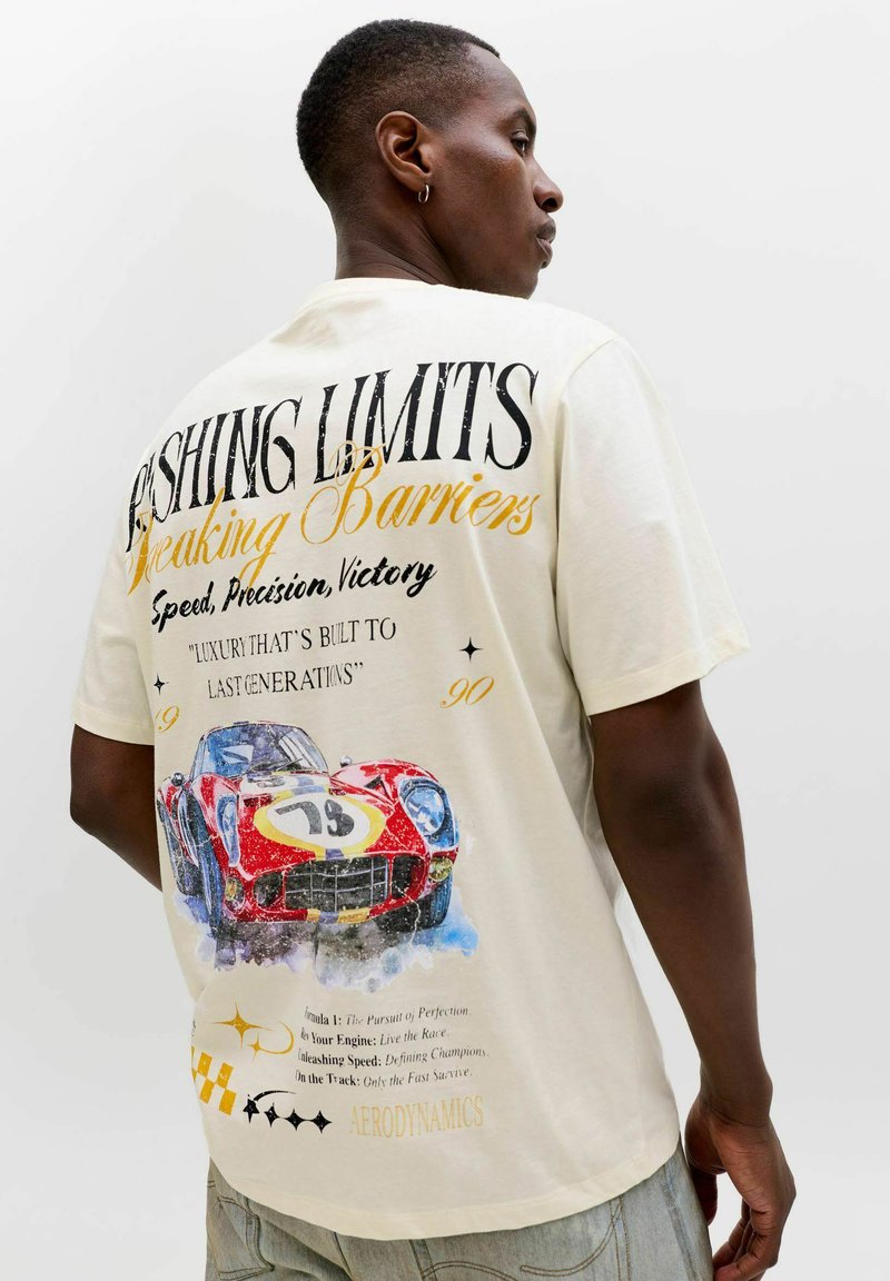 Homme portant un t-shirt crème avec une voiture de course rouge vintage et un texte à thème de course au dos, debout devant un fond blanc.