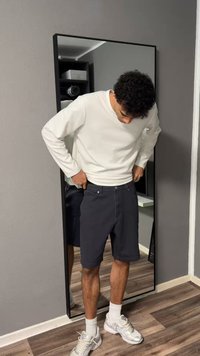 Άνδρας που φοράει λευκό μακρυμάνικο πουκάμισο και μαύρα σορτς, ρυθμίζει τη μέση των σορτς ενώ στέκεται μπροστά σε καθρέφτη ολόκληρου του σώματος σε δωμάτιο.