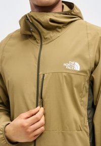 Άνδρας που φοράει μπουφάν North Face ελιάς με τσέπη με φερμουάρ στο στήθος, ρυθμίζοντας το εμπρόσθιο φερμουάρ κοντά στον γιακά.
