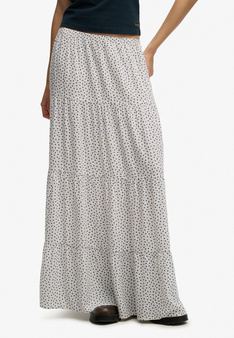 Femme portant une longue jupe blanche à volants avec un petit motif floral bleu et des bottes noires.