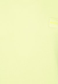 Jasnożółta tkanina z małą naszywaną prostokątną etykietą, na której widnieje słowo "BOSS" w dopasowanym żółtym kolorze.