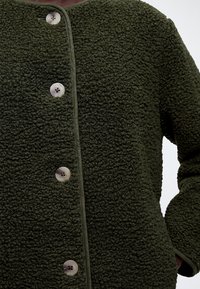 Gros plan d’une veste boutonnée texturée vert foncé avec quatre boutons ronds beige et une bordure lisse le long du devant et des poignets.