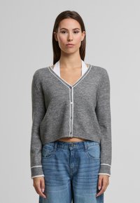 Szürke, puha anyagból készült, rövidített kardigán fehér díszítéssel, V-nyakkal és gombos záródással. Fehér toppal és kék farmerral viselve.