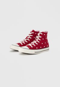 Κόκκινα ψηλά αθλητικά παπούτσια από καμβά, με λευκά σχέδια καρδιών, λευκά κορδόνια και μαύρη ρίγα στη λαστιχένια σόλα.