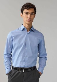 Camicia button-up azzurro chiaro con un sottile motivo a pois, maniche lunghe e colletto a punta, indossata con una cintura nera e pantaloni scuri.