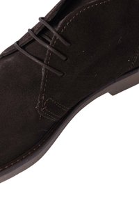 Scarpa in suede marrone con tre fori, cuciture a contrasto e suola in gomma. L'angolo mostra l'altezza della scarpa e la texture dettagliata.