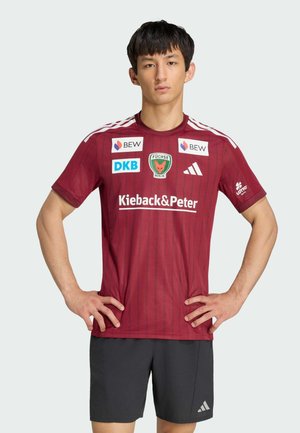 Junger Mann steht mit den Händen in der Hüfte, trägt ein weinrotes Sporttrikot mit Logos und schwarze Shorts vor einfarbigem Hintergrund.