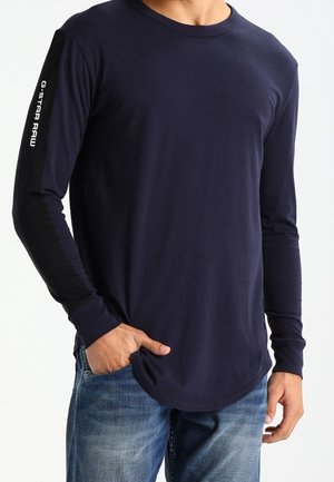 Homme portant une chemise à manches longues bleu marine avec le texte "G-STAR RAW" sur le bras gauche, associé à un jean bleu, debout avec une main dans la poche.