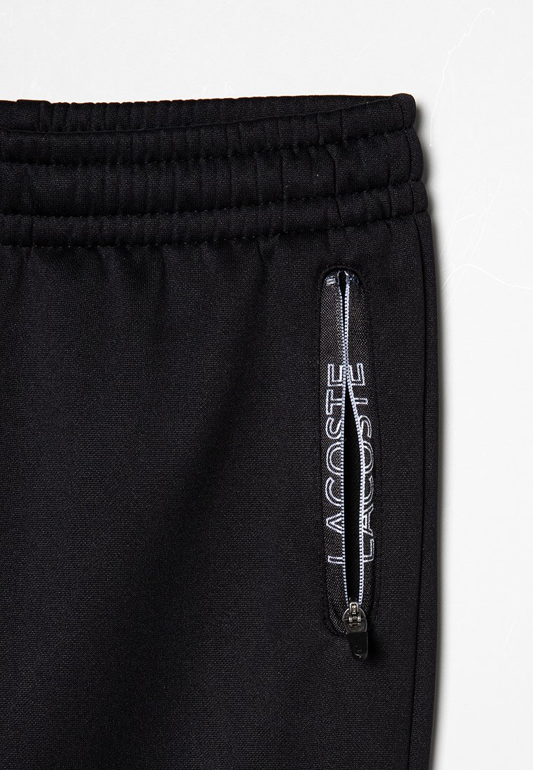 Pantalons de jogging noirs avec taille élastique, poche latérale zippée arborant le logo « Lacoste » en blanc, tissu lisse et coupe fuselée.