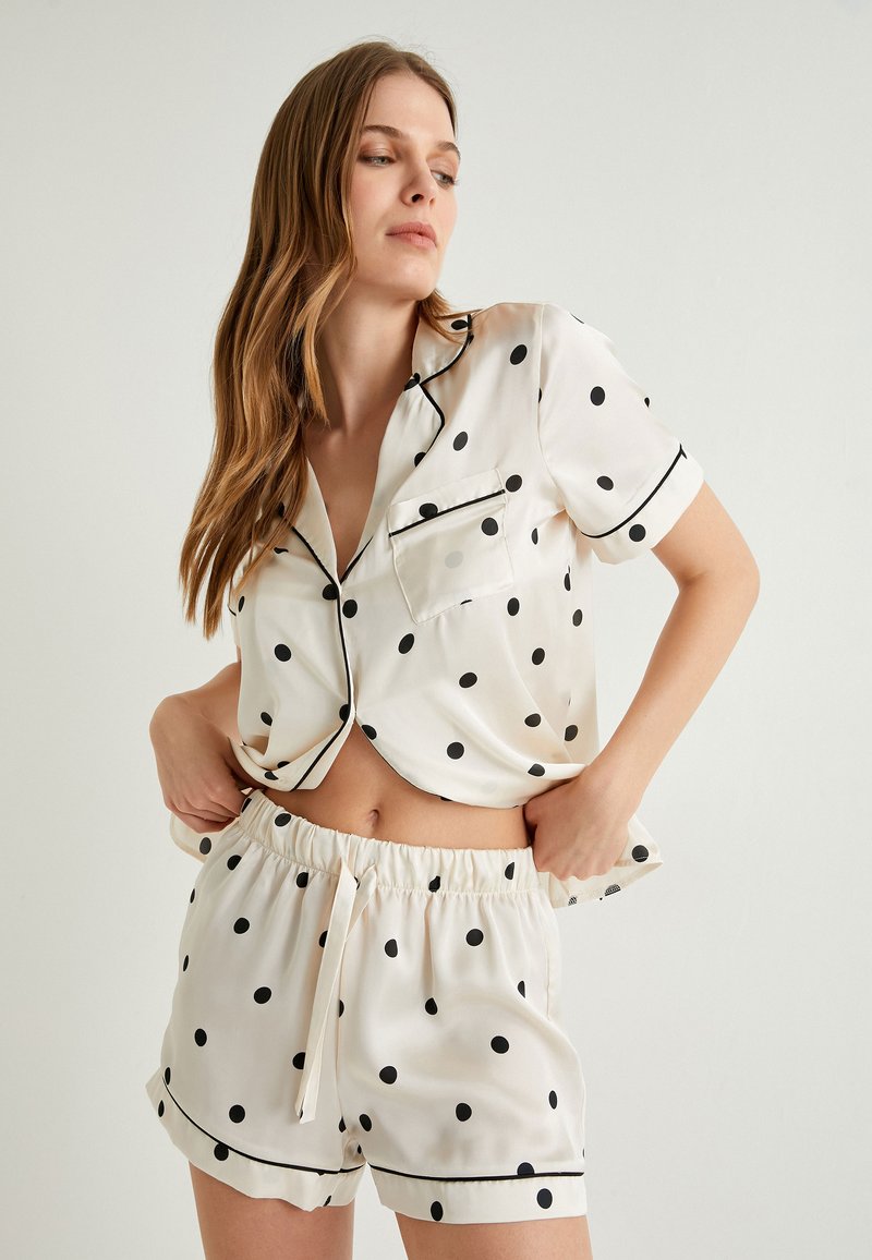 Lyhyet silkkiset pyjamapuvut kermansävyssä, joissa on mustia pilkkuja. Sisältää kauluspaidan, jossa on tasku, ja resorin kanssa varustetut shortsit, joissa on nyöri.