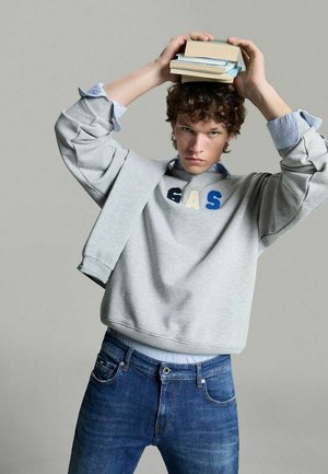 Felpa grigia con scritta "GAS" in blu e crema, indossata sopra una camicia colletto azzurro chiaro, abbinata a jeans blu scuro. Libri impilati tenuti sopra.