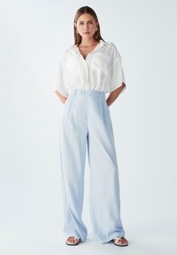 Lichtblauwe wijde broek met hoge taille en een enkele knoopdetail, gecombineerd met een losse witte blouse met twee voorzakken.