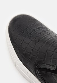Gros plan sur une chaussure noire à enfiler avec un motif en crocodile texturé et une semelle en caoutchouc blanche sur un fond blanc.