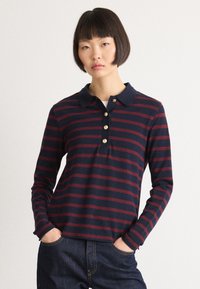 Young woman with short dark hair wearing a navy and burgundy striped long-sleeve polo shirt and dark jeans with hands in pockets.