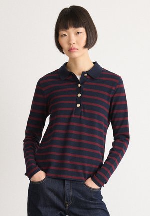 Young woman with short dark hair wearing a navy and burgundy striped long-sleeve polo shirt and dark jeans with hands in pockets.