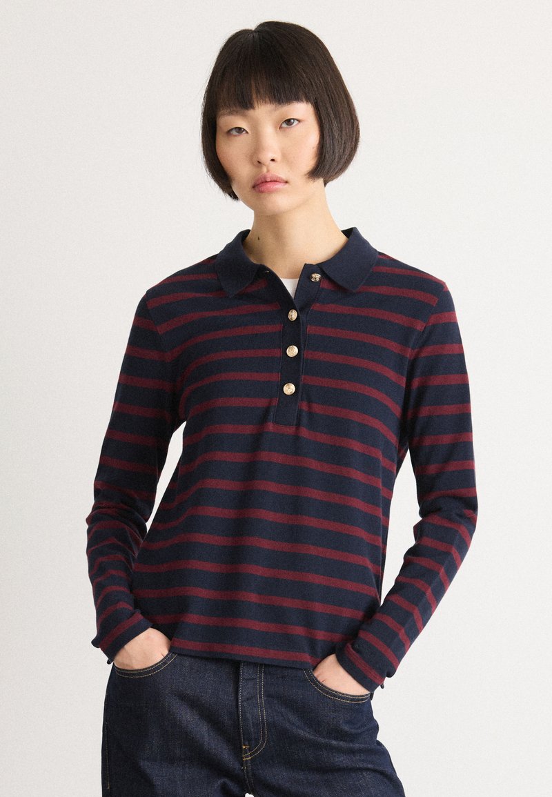 Young woman with short dark hair wearing a navy and burgundy striped long-sleeve polo shirt and dark jeans with hands in pockets.