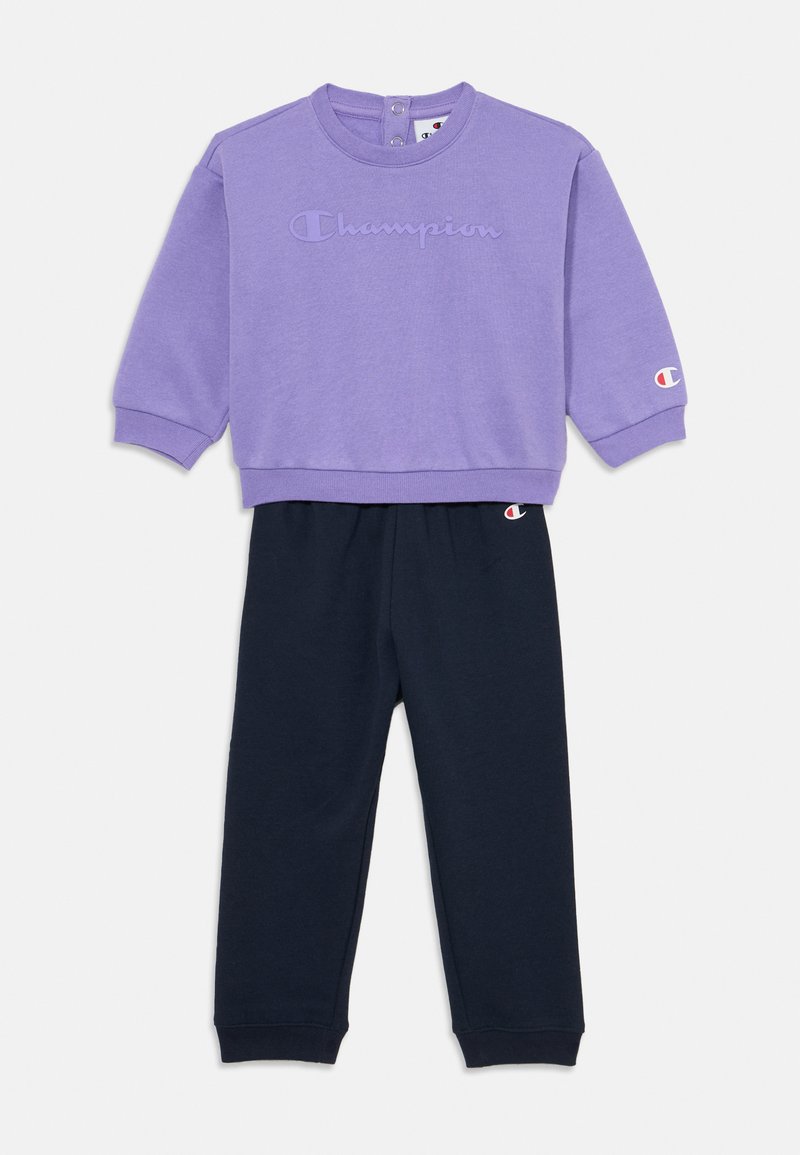 Paarse sweatshirt met reliëf "Champion" logo, gecombineerd met marineblauwe broeken. Het sweatshirt heeft een ronde halslijn en knoopdetails op de rug.