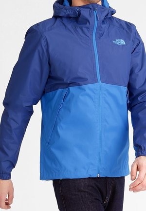 Mężczyzna noszący dwukolorową niebieską kurtkę North Face z przednim zamkiem i kapturem, stojący z ramionami swobodnie opuszczonymi wzdłuż boków.