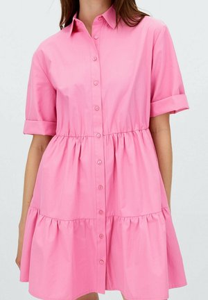 Woman wearing a pink, short-sleeve, button-down dress with gathered tiered skirt and rolled cuffs, standing against a plain background.