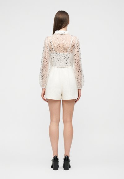 Femme portant un short blanc texturé taille haute et un haut transparent à manches longues avec des pois noirs, vue de dos, debout sur un fond blanc.