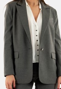 Blazer grigio con vestibilità su misura, rever a punta, due tasche e chiusura con un bottone, abbinato a una camicia bianca con bottoni sotto.