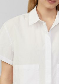 Witte katoenen blouse met korte mouwen, een puntkraag en twee frontzakken. Gladde textuur en een strak ontwerp met een subtiele glans.