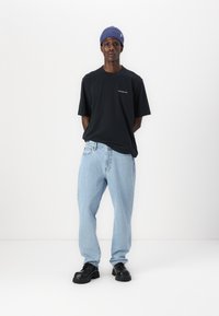 Camiseta negra de manga corta con un pequeño logo blanco, jeans sueltos de color azul claro y zapatos negros gruesos. Gorro de punto azul en la cabeza.