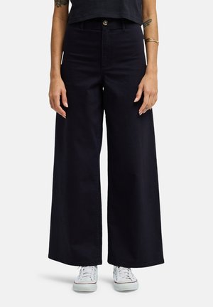 Czarne dżinsy o szerokich nogawkach z wysokim stanem, z przednimi kieszeniami i zapięciem na guziki. Noszone z białymi trampkami.