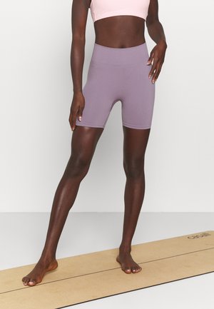 Mujer atlética de pie descalza sobre una esterilla de yoga, vistiendo shorts ajustados color malva y un sujetador deportivo rosa claro frente a una pared lisa.