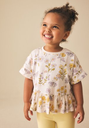 Lächelndes junges Mädchen mit lockigem Haar, das eine geblümte, mit Rüschen verzierte Bluse und gelbe Hose vor beigem Hintergrund trägt.