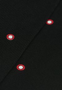Tejido negro acanalado con detalles bordados circulares en rojo y blanco. Dos secciones distintas con patrones contrastantes.