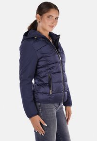 Giacca puffer blu navy con un design aderente, cappuccio e due tasche frontali con zip. Presenta una texture liscia e lucida con dettagli in hardware dorato.