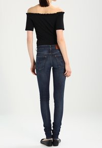 Top negro de corte asimétrico con cuello fruncido, combinado con jeans ajustados azul oscuro, que presentan una costura del bolsillo trasero minimalista y una textura suave.