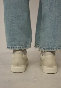 Baskets en cuir crème à surface texturée, posées sur un sol gris clair, associées à un jean large en denim bleu légèrement effiloché.