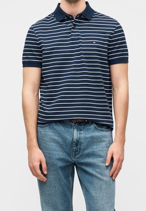 Homme portant un polo à rayures horizontales bleu marine et blanc à manches courtes et un jean bleu, debout devant un fond uni.