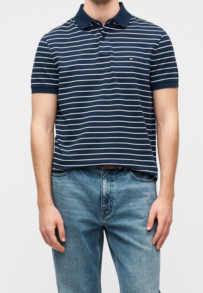 Homme portant un polo à rayures horizontales bleu marine et blanc à manches courtes et un jean bleu, debout devant un fond uni.