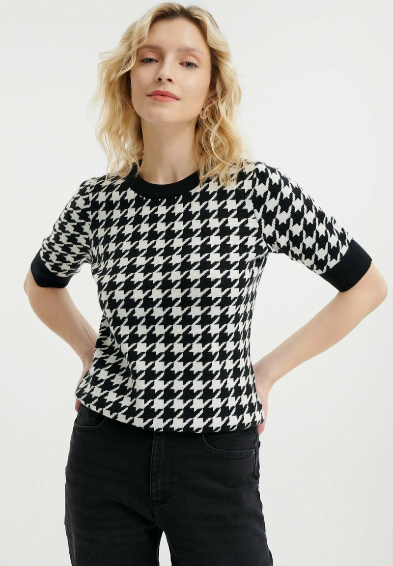 Top s černobílým károvaným vzorem houndstooth s krátkým rukávem a černým kulatým výstřihem, vyrobený z jemného pleteného materiálu.