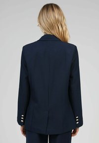 Blazer ajusté bleu marine avec un col cranté, une fente simple et des accents de boutons dorés sur les manches. Texture de tissu lisse.