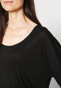 Kobieta o długich, ciemnych włosach, ubrana w luźną czarną bluzkę z szerokim dekoltem odsłaniającym jej obojczyk i szyję.