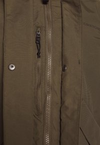 Gros plan sur le devant d'une veste marron montrant une fermeture éclair, une petite poche verticale zippée avec languette, et des boutons-pression.