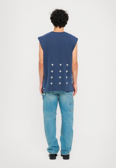 Cămașă fără mâneci de culoare bleumarin, cu guler rotund și semne albe plus imprimate pe spate, asortată cu pantaloni cargo de un bleu deschis și pantofi negri.