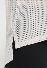 Lichte, ademende mesh-top in lichtroze met kleine zwarte perforaties. Heeft zijsplitten aan de onderrand en een relaxte pasvorm.