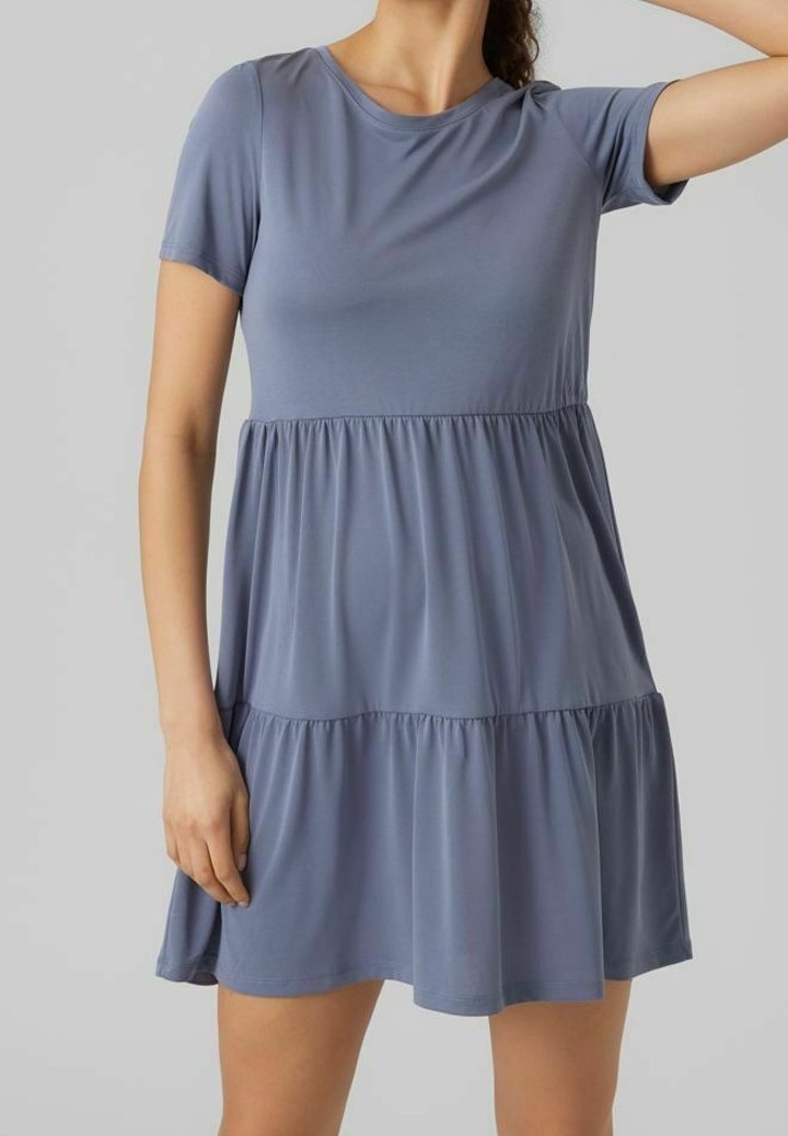 Femme portant une robe bleu clair à manches courtes et à étages, debout devant un fond gris uni.