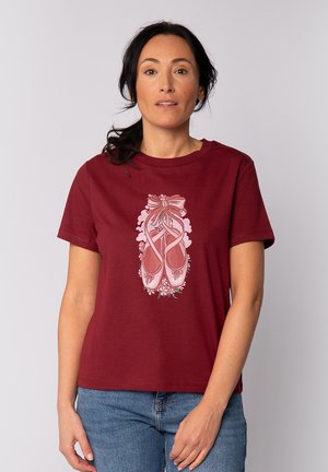 Vrouw draagt een bordeauxrood T-shirt met een ontwerp van roze balletschoenen en bloemen, gecombineerd met een blauwe spijkerbroek, staande tegen een effen achtergrond.