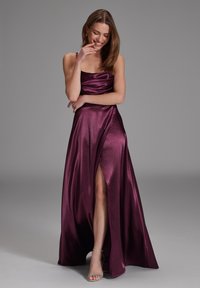 Robe longue sans manches en satin bordeaux riche avec un corsage ajusté et une fente sur le côté. Elle présente une texture lisse et un léger éclat.