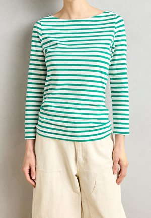 Femme portant un haut à manches longues à rayures horizontales vertes et blanches et un pantalon beige à jambes larges sur un fond uni.