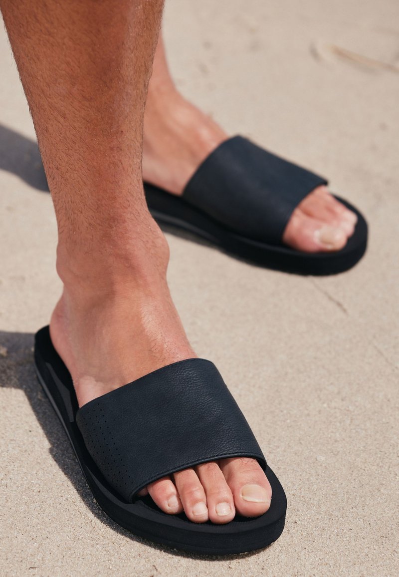 Zwarte leren slidesandalen met een brede band, een gestructureerd bovenwerk en een dikke zwarte zool, uitgestald op een zanderige ondergrond.