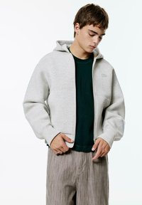 Sivá mikina na zips vyrobená z mäkkého materiálu, s kapucňou a manžetami na rukávoch. Pod ňou je vidieť tmavozelené tričko. Pásové nohavice s béžovými pruhmi.