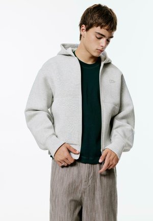 Hanorac zip-up gri, realizat dintr-un material moale, având glugă și mâneci cu manșete. Sub acesta, se vede un tricou de culoare verde închis. Pantaloni bej cu dungi.